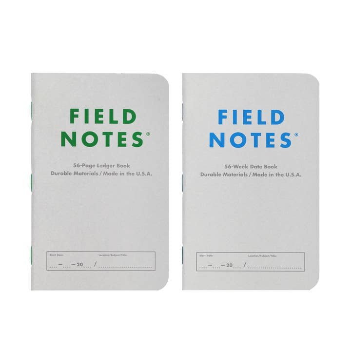 Two white Field Notes memo books stand upright: a 56-page Ledger Log Book and the 56-Week Date Book by Field Notes, both from the Index Edition with simple covers and colored text.