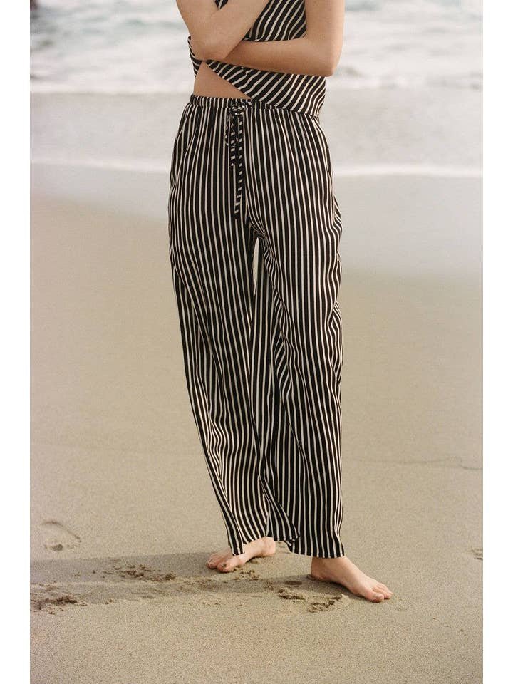 A person stands barefoot on the sand by the ocean, arms crossed, wearing A. Ren’s black and white striped Amina Pants with a matching top.