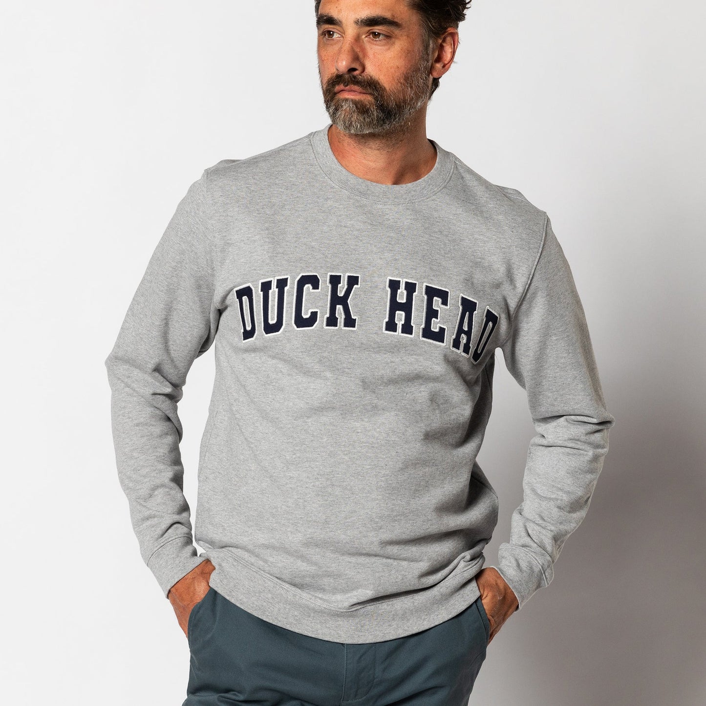 Man wearing the Duck Head Applique Crewneck Sweatshirt—a light gray cotton crewneck with a bold black "DUCK HEAD" appliqué logo on the front—stands hands in pockets against a plain background.