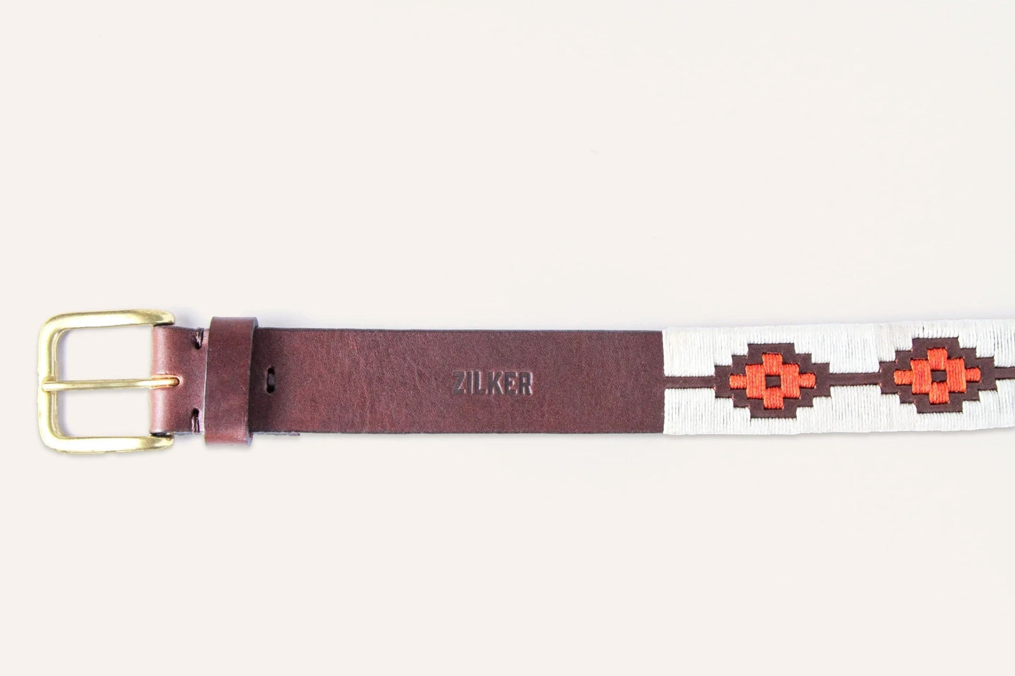 The ATX Dark by Zilker Belts is a brown Argentine leather belt with a gold buckle, hand-stitched white and red geometric pattern, and "ZILKER" stamped on it.