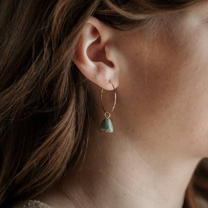 Close-up of a woman's ear adorned with the Aurora Hoops by CommonForm—a handmade brass hoop earring featuring a green stone pendant; brown hair partially visible.
