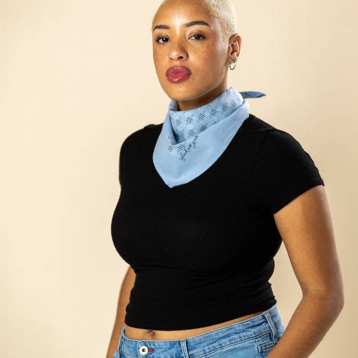 A person with short blond hair, in a black fitted top and blue jeans, wears the "Fuck All Y'All" Bandana by Jenni Earle around their neck, standing against a beige background.