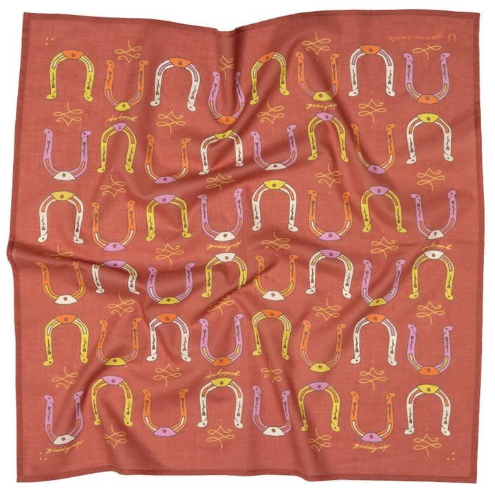 The Jenni Earle "Giddy Up" Bandana features a red background with multicolored horseshoes and yellow abstract shapes. Its unique folded look makes it an uplifting accessory or thoughtful encouragement gift.