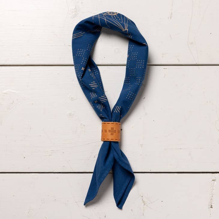 The "Hell Yeah" Bandana by Jenni Earle, featuring a blue pattern and made in the USA, is looped through a light brown wooden ring and laid flat on a white wooden surface.