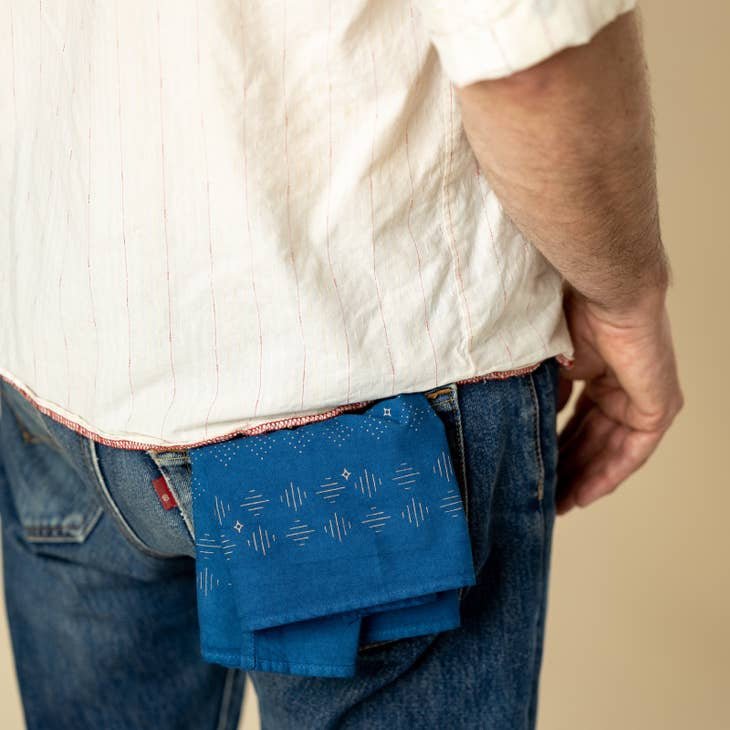A person in jeans and a white shirt has a "Hell Yeah" bandana by Jenni Earle, made in the USA, featuring a blue pattern, sticking out of their back pocket.