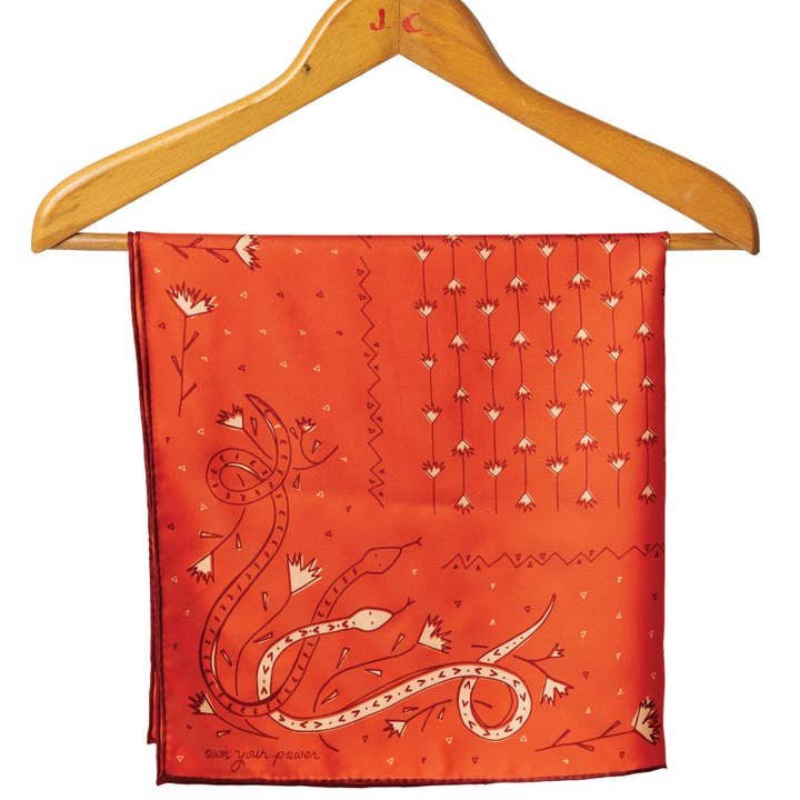 The “Own Your Power Silk” Bandana by Jenni Earle features a red snakes, flowers, and geometric print—draped on a wooden hanger. Perfect as a stylish accessory or unique holiday gift.