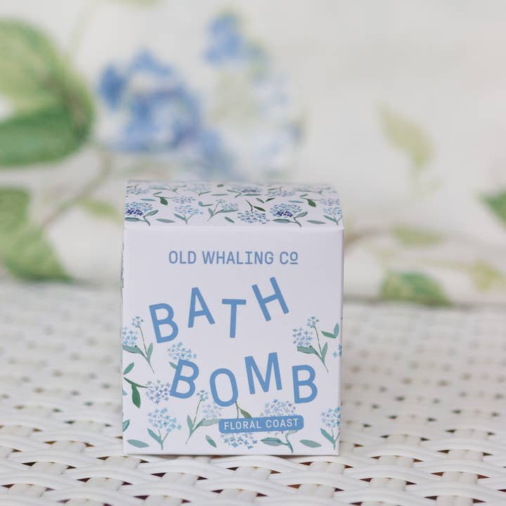 A box labeled "Bath Bomb | Floral Coast | Old Whaling Co." with a floral design and hydrangea scent rests on a white woven surface.