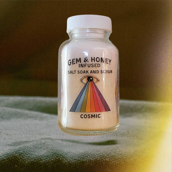 A clear jar of Wild Yonder Botanicals Bath Soak | Cosmic sits on a gray surface with a brown background, featuring a rainbow eye design—ideal for anyone seeking a unique botanical bath experience.
