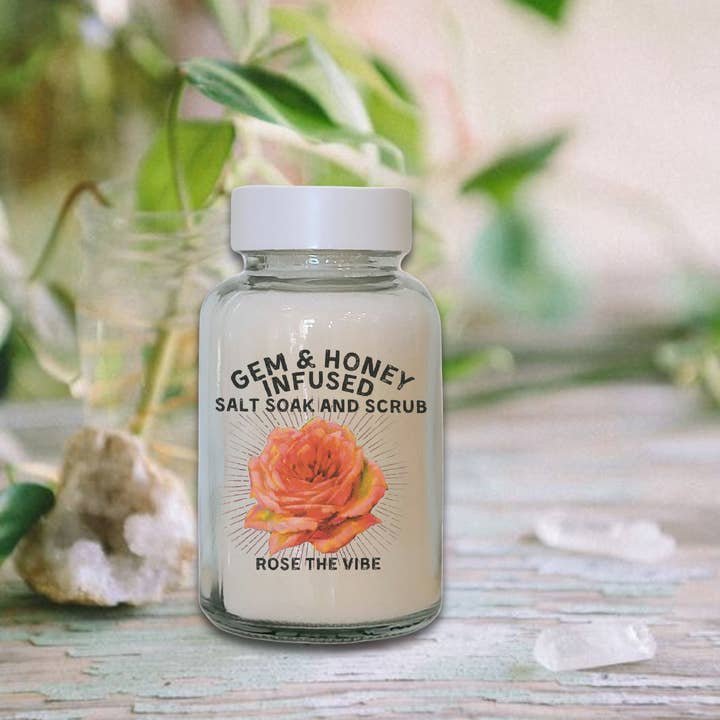 A clear jar labeled "Bath Soak | Rose the Vibe | Wild Yonder Botanicals" with an orange rose design sits on a table amid crystals and greenery—ideal for a relaxing rose bath soak by Wild Yonder Botanicals.