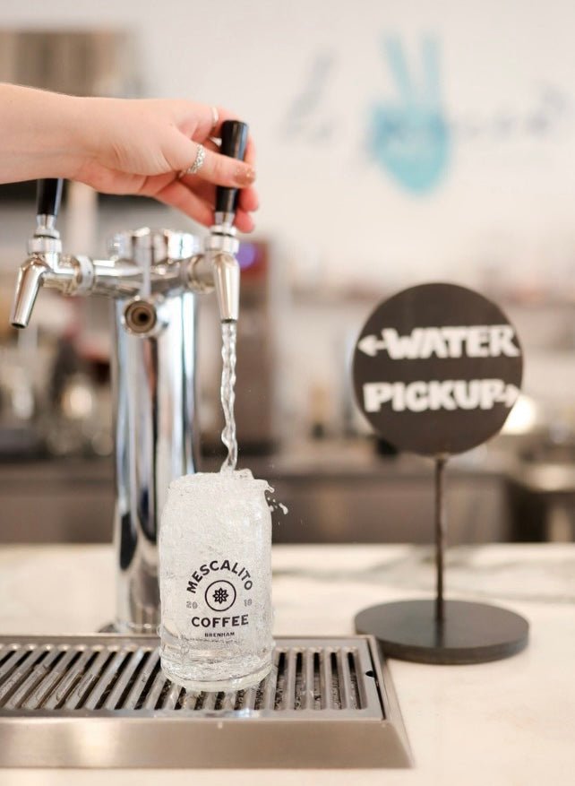 A hand fills a Beer Can Glass from Mescalito Coffee with water at a "WATER PICKUP" station, making it ideal for any cold beverage.