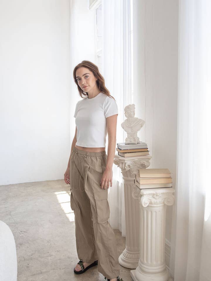 A woman stands indoors next to a column with stacked books and a bust, wearing the Billie Pant by Things Between—a beige parachute style with an adjustable waist—paired with a white t-shirt.
