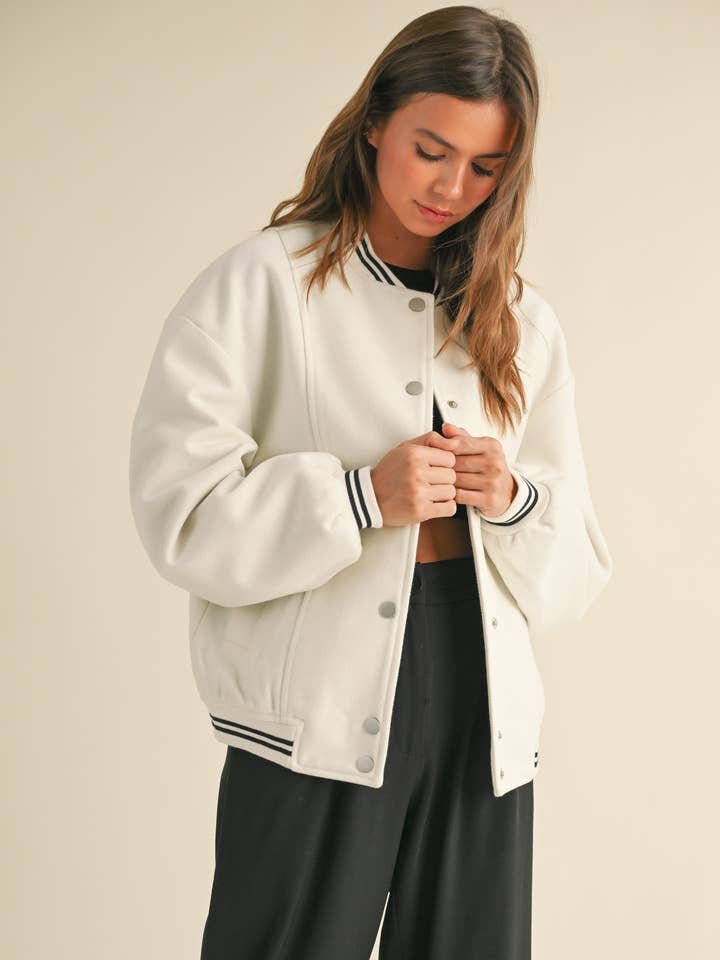 A woman stands looking down, buttoning the Miou Muse Bomber Jacket—white with black striped cuffs and collar—over a black top and pants.