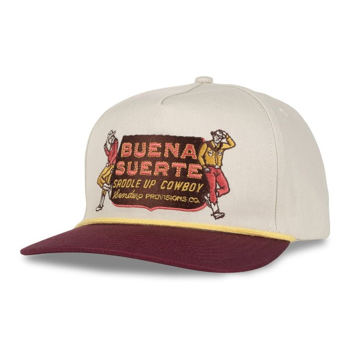 The Buena Suerte Hat by Sendero Provisions Co. is a beige and maroon embroidered snapback with a curved visor, featuring a patch reading "BUENA SUERTE SADDLE UP COWBOY" and cowboy illustrations on each side.