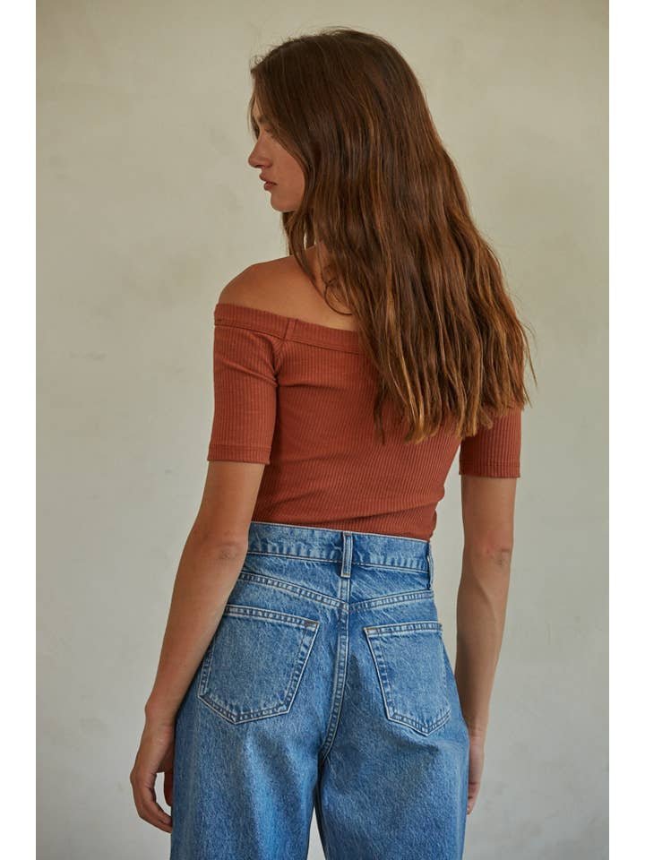 A woman with long brown hair wears the By Together Calla Top, a rust-colored ribbed knit off-the-shoulder style, paired with high-waisted blue jeans, standing with her back to the camera against a plain background.