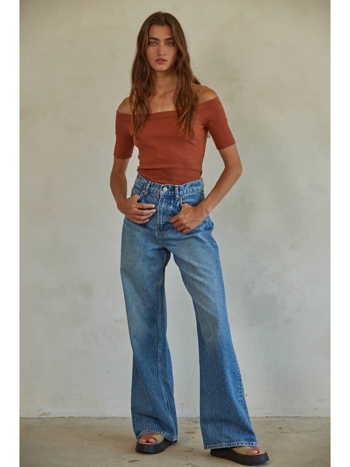 A person stands against a plain wall wearing the rust Calla Top by By Together, high-waisted blue wide-leg jeans, and black slide sandals.