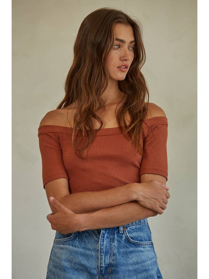 A woman with long brown hair stands with her arms crossed, looking to the side, wearing the Calla Top by By Together—an off-the-shoulder ribbed knit in rust—paired with blue jeans.