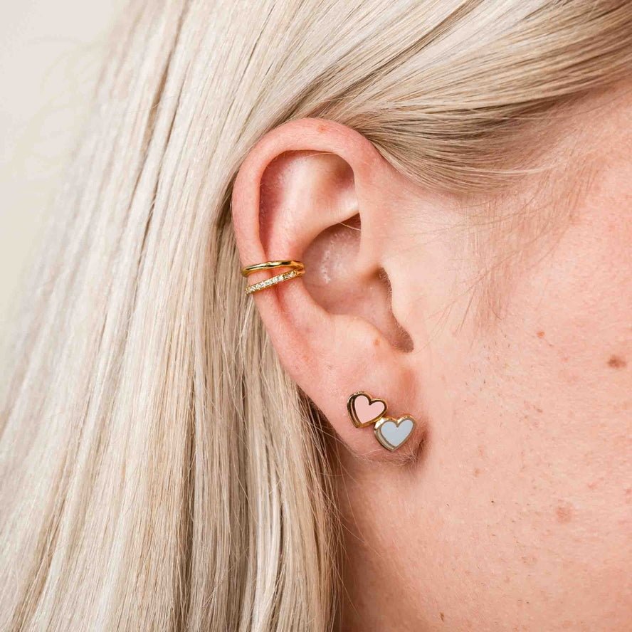 A close-up of a person’s ear with two gold hoops on the upper ear and two Larissa Loden Candy Heart Enamel Studs, hypoallergenic gold-plated heart earrings, in the earlobe.