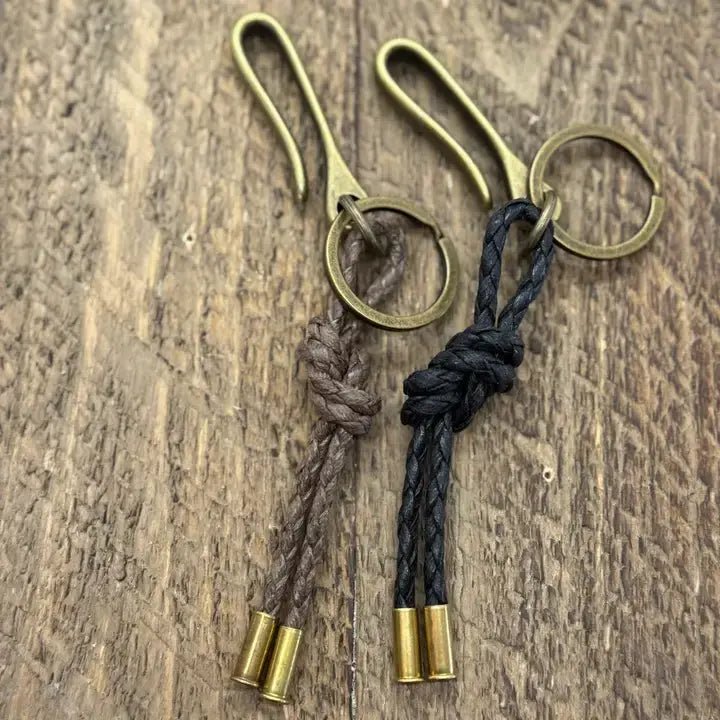 Two Tres Cuervos Canvas Ocho Keychains—each with brass hardware and braided leather loops in brown and black, figure 8 knot, finished with two brass caps—rest on a wooden surface.