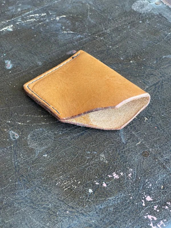 A small, tan leather handmade Card Wallet by James Foreman with visible stitching rests on a worn, dark textured surface.