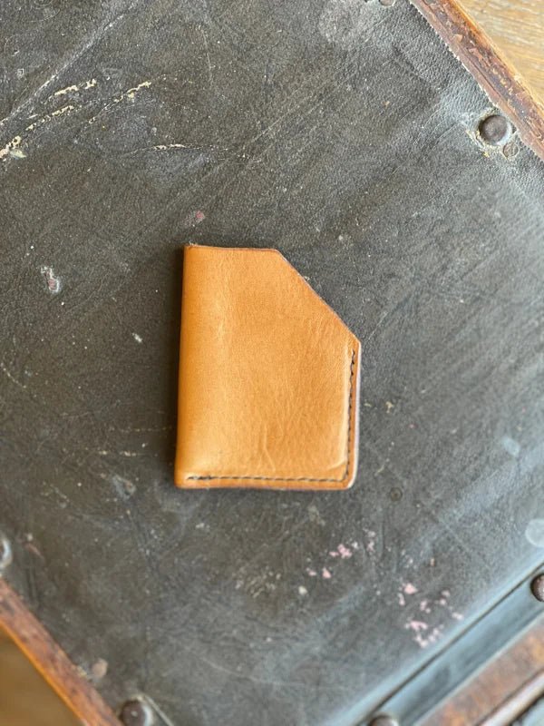 A James Foreman Card Wallet in tan leather with a handmade, angled-top design rests on a worn, dark surface marked by scratches and rivets.
