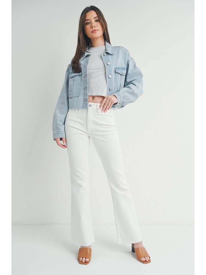 A woman stands against a white background wearing the JBD Cargo Jacket in light denim, styled with a gray crop top, white flared pants, and tan heeled sandals for an effortless Spring/Summer look.
