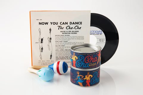 A cha-cha dance instruction record, its cover showing dance steps, a pair of maracas, Beautiful Briny Sea’s Cha Cha Delish Seasoning, and a tin labeled "Cha Cha Chilis" are arranged together against a white background.