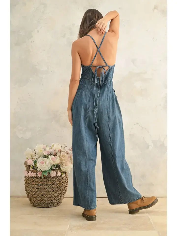 A person stands facing away, wearing the Miou Muse Chambray Fabric Jumpsuit with crisscross straps and brown boots beside a woven basket of flowers.