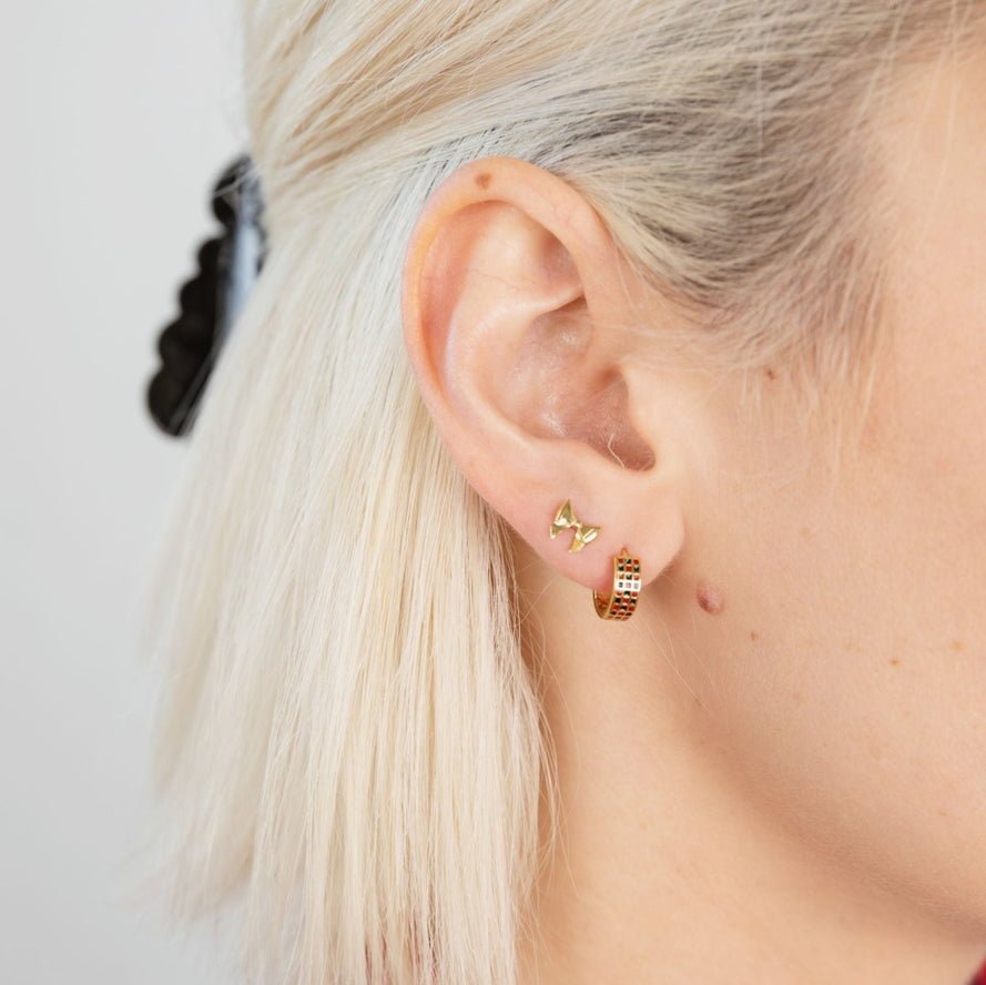 Close-up of an ear with two earrings: a small 18k gold plated butterfly stud and Larissa Loden’s Checker Huggie Hoops—a gold hoop with a checkered pattern. The person has light blonde hair pulled back with a black hair clip.
