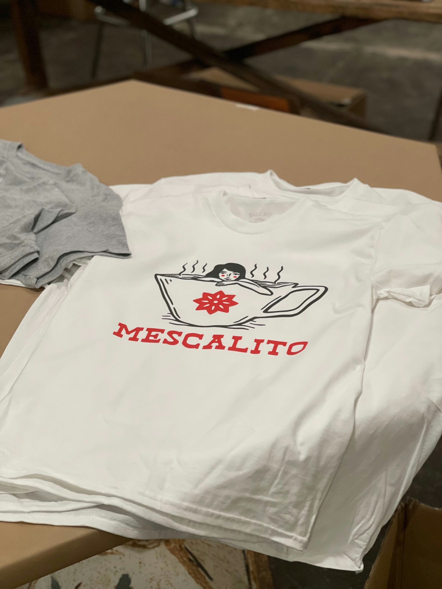 The Coffee Jacuzzi Girl t-shirt by Mescalito Coffee features a steaming cup with a face and "MESCALITO" in red, inspired by Mescalito Coffee vibes. A gray t-shirt is partially visible on the left.