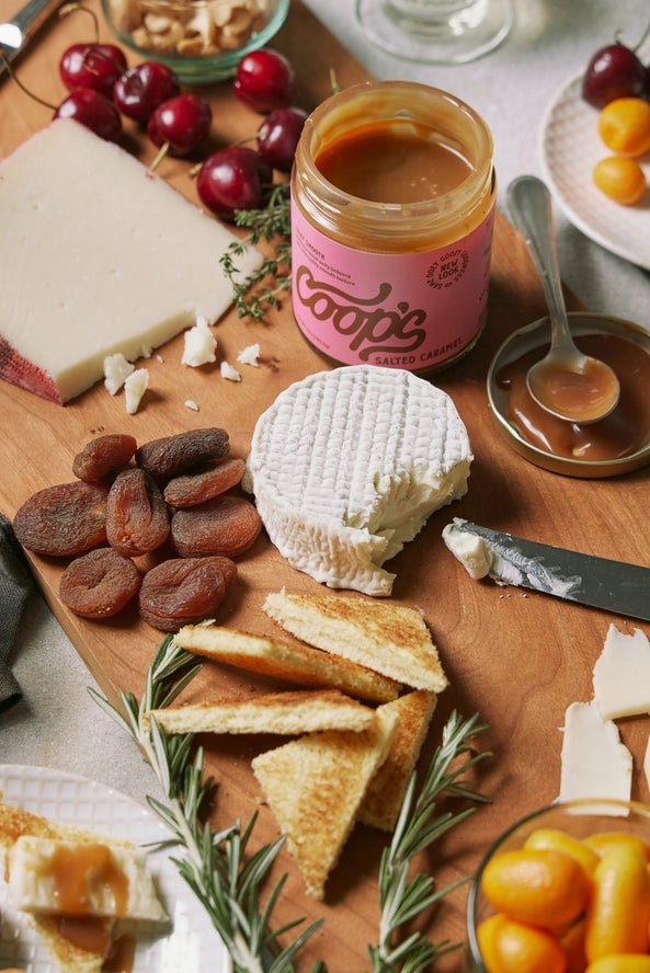 A wooden board with cheese, dried apricots, toasted bread, cherries, nuts, and a jar of Coop's | Salted Caramel served with a knife and spoon.