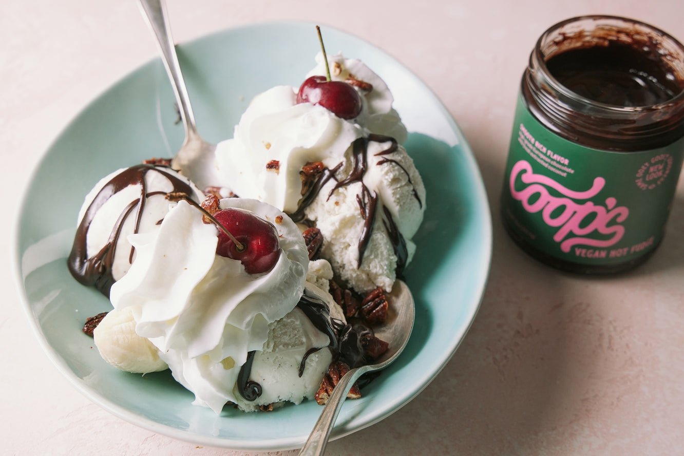 A bowl of banana split topped with whipped cream, cherries, European chocolate sauce, and pecans, served with Coop's Vegan Hot Chocolate by Coop's on the side. Perfect as a dairy-free ice cream topping or for any sweet treat!.