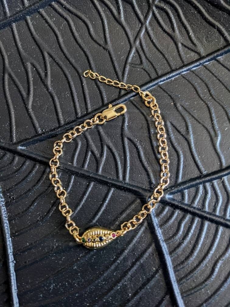 The Coquille D'or Bracelet by Minh Atelier, an 18k gold plated shell charm bracelet with a cowrie shell, is displayed on a textured black surface with a geometric pattern.