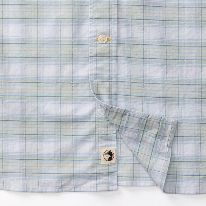 Close-up of Duck Head’s Cotton Oxford Sport Shirt in Raney Plaid, featuring light blue and beige checks, detailed buttons and stitching, plus a small bird patch near the hem.