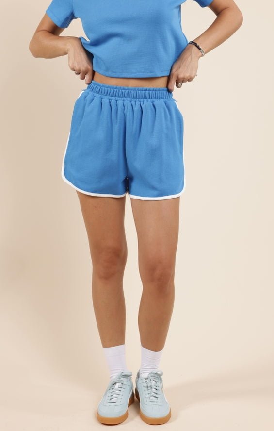Model showcases Courtside Piped Dolphin Shorts with pockets by Sadie And Sage, paired with a blue cropped cotton-poly tee, white socks, and blue sneakers against a beige background.
