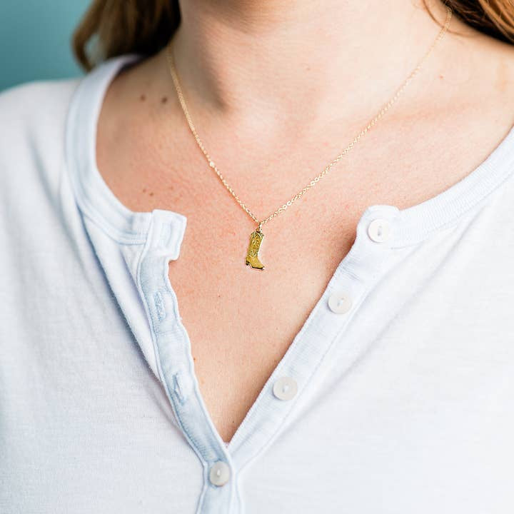 A person wearing a white buttoned shirt and the Cowgirl Boot Necklace by Ornamental Things, which features a gold filled chain and a gold boot-shaped pendant.