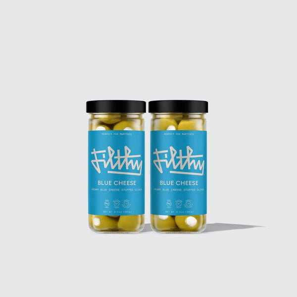 Two jars of Filthy Creamy Blue Cheese Stuffed Olives sit side by side on a plain light gray background—the perfect bold martini garnish, filled with smooth Wisconsin blue cheese.