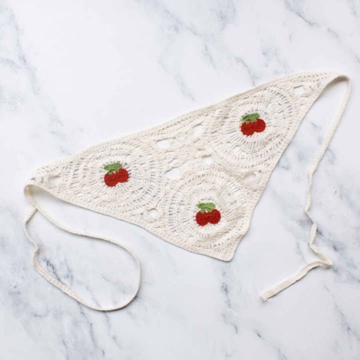 A white, crocheted triangle bikini top with three red strawberry accents is laid flat on marble—pair it with the Tiepology Crochet Triangle Headscarf for versatile styling.