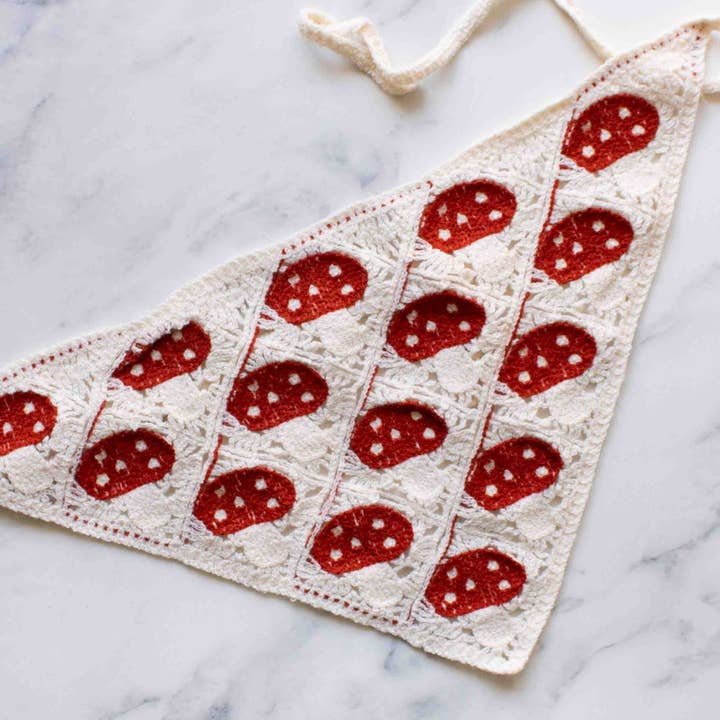 The Tiepology Crochet Triangle Headscarf features a white border and rows of red and white mushroom motifs, shown on a marble surface—an elegant accessory to add charm to any outfit.