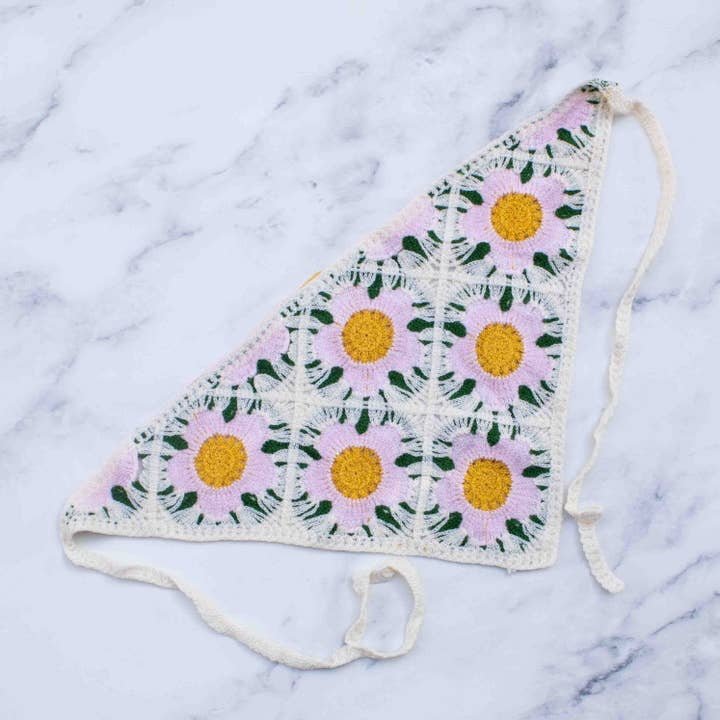 The Crochet Triangle Headscarf by Tiepology features pink and yellow floral crochet, white tie strings, and a chic triangular shape—perfect as a versatile accessory or elegant headscarf. Displayed on a white marble surface.