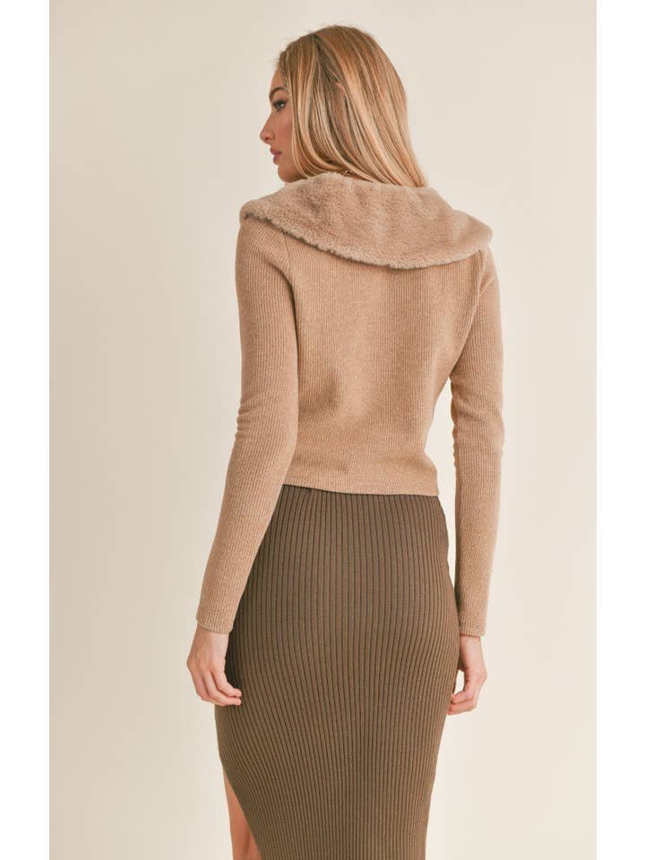 A woman is shown from the back wearing the Daria Vegan Fur Cardi by Sage the Label, a tan long-sleeve top with a faux fur collar, paired with a high-waisted, ribbed olive green spandex-blend pencil skirt.
