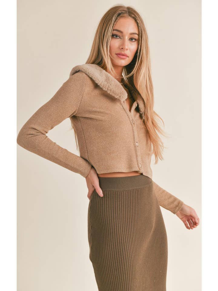 A woman wears the Daria Vegan Fur Cardi by Sage the Label—a tan ribbed long-sleeve crop top with a faux fur collar—paired with a high-waisted brown knit skirt, all in cozy spandex blend, standing against a neutral background.