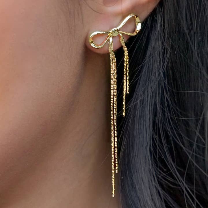 A close-up of a person wearing Peter and June's Disco Bow Stud: 8k gold-plated, bow-shaped earrings with long, thin waterproof dangling strands that beautifully frame the face.