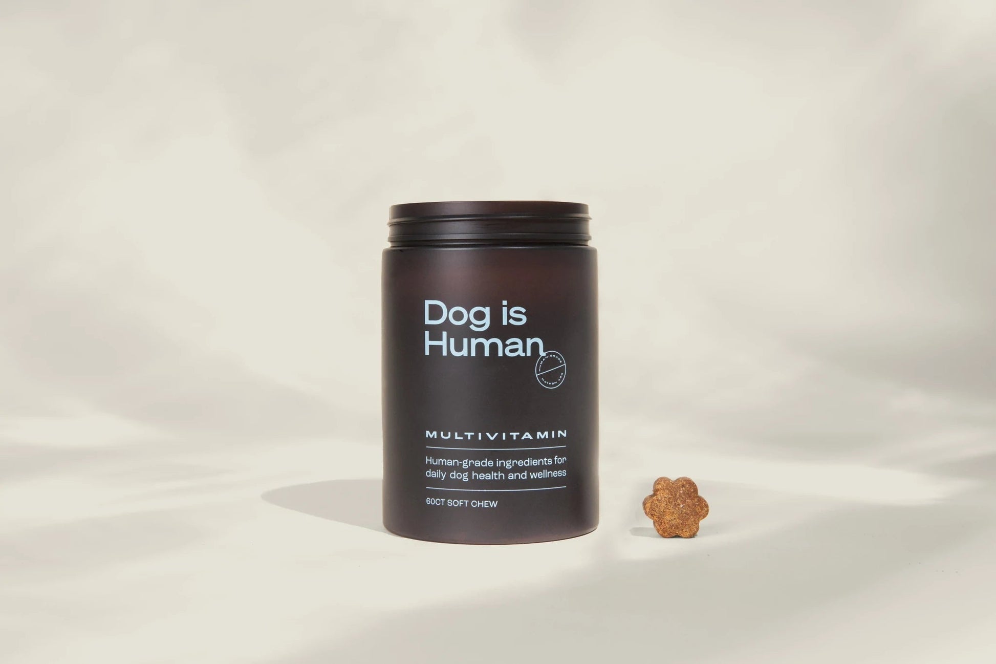 A dark container labeled "DM-01™ Daily Multivitamin | Dog is Human" sits beside a small, paw-shaped probiotic chew for hip and joint support, both displayed against a neutral background.