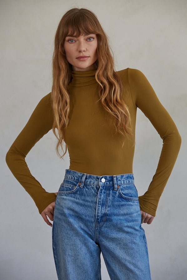 A woman with long wavy hair wears the mustard yellow Done For Me Bodysuit by By Together and high-waisted blue jeans, standing with hands on hips against a neutral background.