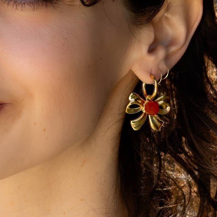 Close-up of a person wearing Peter and June's Knotty Hoops 18k gold plated flower-shaped earring with a red center, shown on their left ear against brown curly hair.