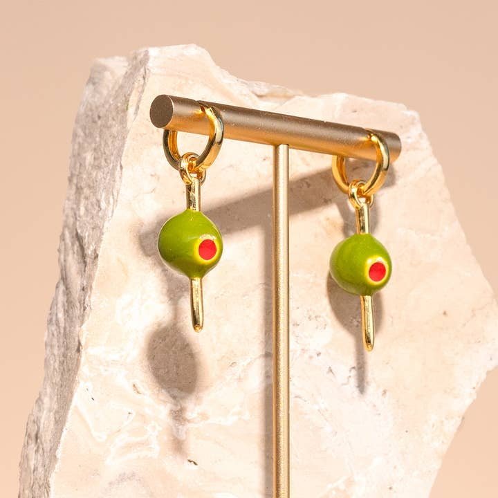 The Peter and June Make it Dirty Hoops are 18k gold plated earrings with green olive-shaped charms, red pimento details, and gold toothpicks, shown on a stone stand.