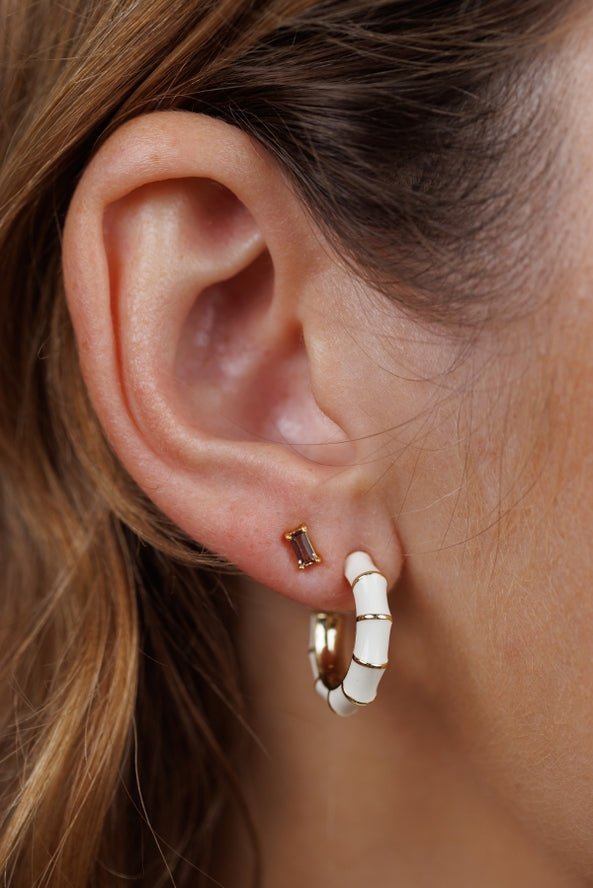 Close-up of a person's ear wearing JaxKelly Vintage Hoop Earrings—stylish, hypoallergenic white and gold enamel hoops paired with a small rectangular stud.