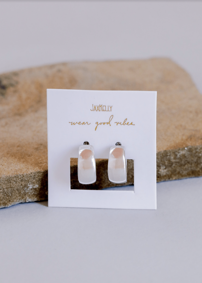 A pair of wide hoop earrings by JaxKelly, perfect for making a statement, displayed on a branded card with "JaxKelly wear good vibes" text, set against a tan stone background.