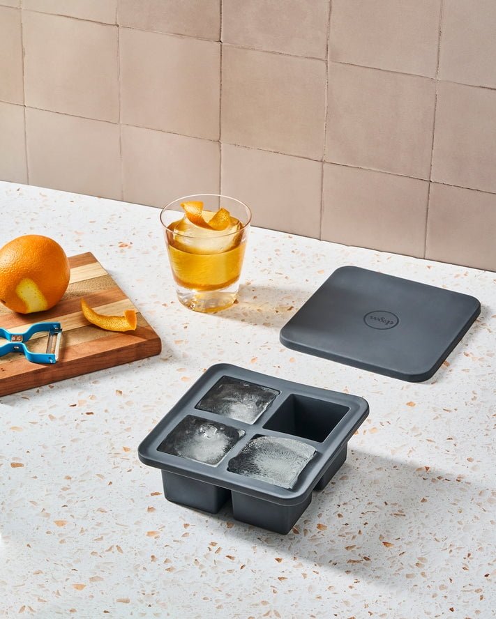 The W&P Extra Large Ice Cube Tray with lid creates oversized cubes. Next to it: a drink garnished with orange peel, a cutting board, an orange, and a peeler—all arranged on a terrazzo countertop.