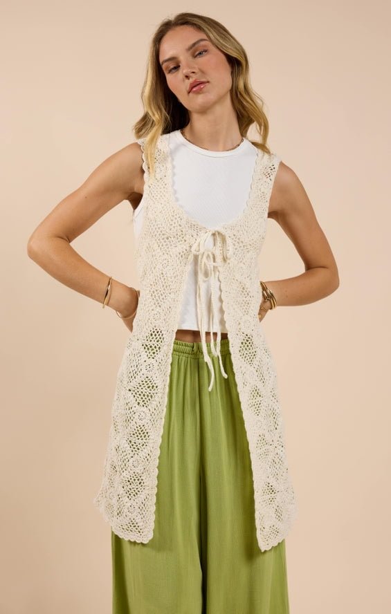 Woman wearing the Fell For You Crochet Front Tie Cardigan by Sadie And Sage over a white sleeveless top and light green pants, standing against a beige background. Stylish choice for women looking for small-size fashion.